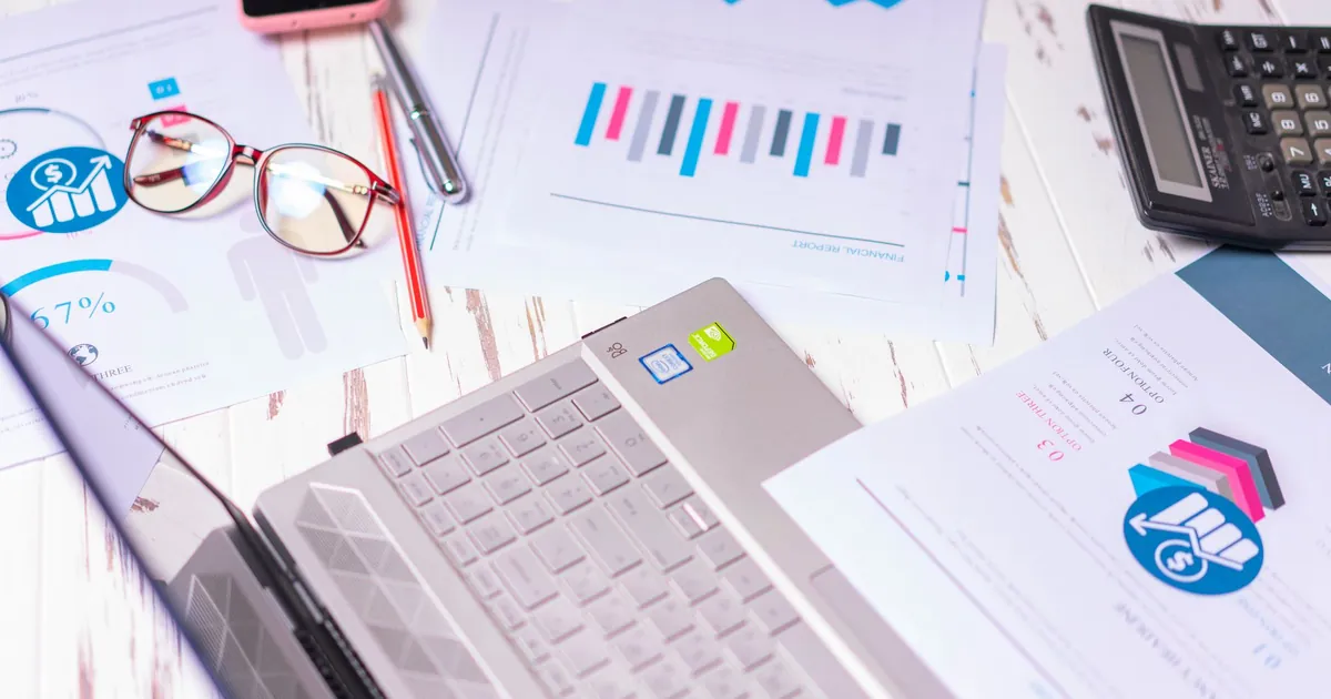 A cluttered desk with a laptop, financial reports, calculator, and glasses indicating a busy work environment.