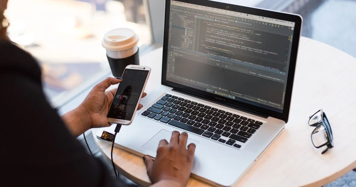 Mobile app developer coding on a laptop with a phone beside it.